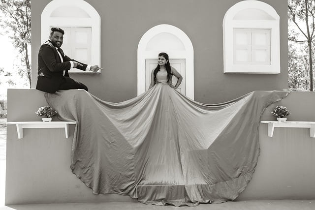 "Black and white pre-wedding photo of a bride standing and groom sitting .The groom, dressed in a coat and pants, is pointing both the finger at bride's gown, while the bride has one hand on her waist. They are posed in front of a background featuring a door in the center with two windows on either side."