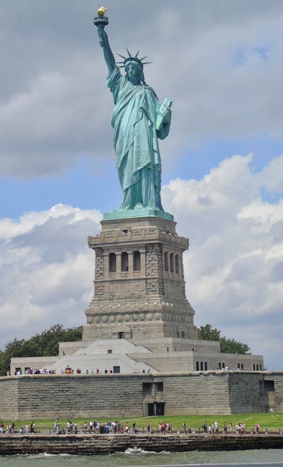 "View of the Statue of Liberty standing majestically on Liberty Island, holding a torch high in her right hand, with a clear blue sky and the Manhattan skyline visible in the background."